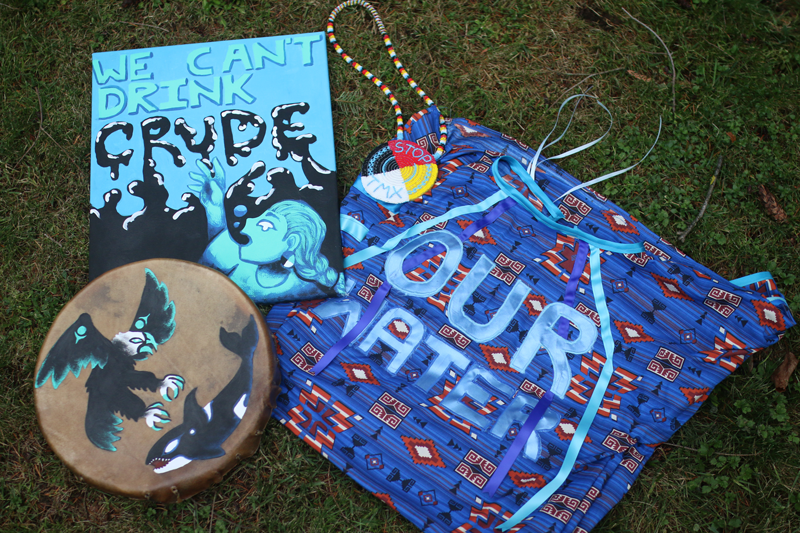 Four items that make the uniform of an Indigenous Environment Protector. These are a ribbon dress with the words “Our Water” on the front, a red, black, yellow and white beaded medallion that says “Stop TMX”, an elk and cedar drum with a Thunderbird and Orca painted on top, and a blue and black protest sign with the words “We Can’t Drink Crude.”